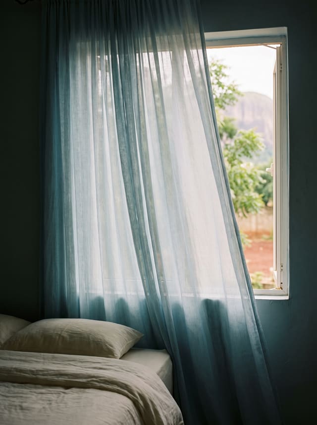 A mist-blue curtain moving at a bedroom window in soft morning light